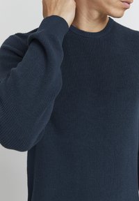 Pull en tricot côtelé bleu marine avec des manches longues et un col rond. La texture est douce avec des détails de couture visibles.
