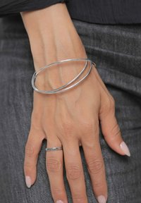 Braccialetto rigido in metallo argentato con due anelli intrecciati, superficie liscia, indossato su una mano che mostra un sottile anello d'argento e uno smalto per unghie neutro.