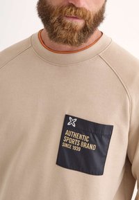 Sudadera beige con un cuello de canalé, que presenta un bolsillo de parche negro. El bolsillo incluye un texto en amarillo: "MARCA DEPORTIVA AUTÉNTICA DESDE 1939."