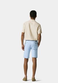 Beige Kurzarmhemd kombiniert mit hellblauen Shorts. Auffällige Details sind die Nähte und zwei Gesäßtaschen. Getragen mit braunen Schuhen.