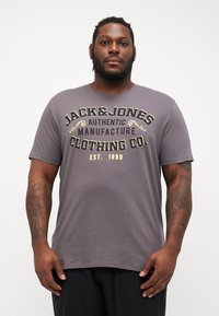 Pelēka kokvilnas T-krekls ar izteiksmīgu melnu un zelta grafiku, kurā ir uzraksts "JACK&JONES AUTHENTIC MANUFACTURE CLOTHING CO. EST. 1990."