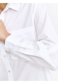 Biała koszula zapinana na guziki, wykonana z gładkiego materiału, z kieszonką na naprasce, długimi rękawami z mankietami na guziki oraz miękką teksturą.