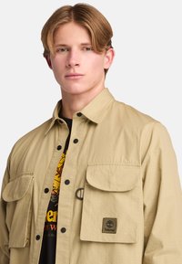 Tan utility shirt van lichtgewicht stof, met twee voorzakken, knoopsluitingen en een logo patch op de borst.