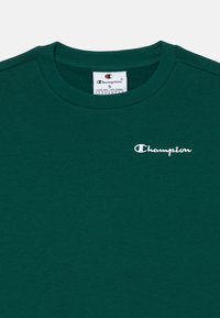 T-shirt in cotone verde con colletto rotondo, caratterizzato da un piccolo logo bianco "Champion" sul lato sinistro del petto. Design semplice e sobrio.