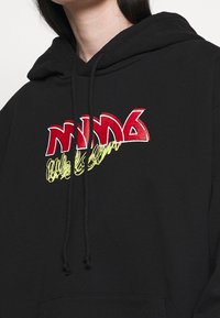 Svart hoodie med djärv röd och gul broderad text. Texten har en lekfull design, kompletterad med svarta dragsko.