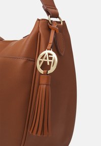 Sac à main en cuir marron avec une étiquette circulaire dorée portant les initiales "AH" et un long pompon en cuir attaché à la sangle.