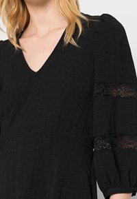Vestido negro con escote en V, tejido texturizado y mangas abullonadas. Acentos de encaje en las mangas añaden un detalle decorativo.