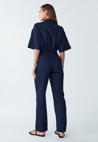 Marineblauwe jumpsuit met korte mouwen, elastische tailleband en rechte pijpen. Soepele textuur zonder zichtbare patronen, gedragen met platte sandalen.