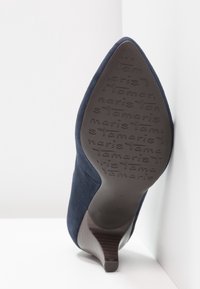 Chaussure à talon haut en daim texturé bleu marine avec un bout pointu et un talon fin. La semelle présente des détails de la marque en relief.
