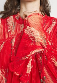 Punainen, laskostettu paita kukkakuosisena, jossa on rypytettyjä, joustavia hihoja ja herkkä narupitsi. Läpinäkyvä, tekstuuriainen kangas.