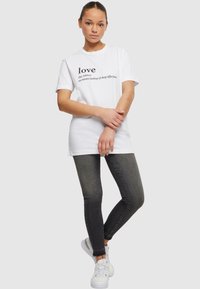 T-shirt blanc à manches courtes comportant du texte noir avec le mot "amour" et sa définition. Porté avec un jean sombre et des baskets blanches.