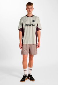Graues gestreiftes Sporttrikot mit kastanienbraunen Akzenten und "SWARM"-Logo, kombiniert mit hellbraunen Shorts und schwarzen Sneakers, dazu weiße gestreifte Socken.