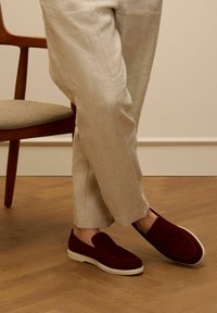 Burgundy suede slip-on shoes with a white sole, worn with light beige linen trousers, on a herringbone wooden floor.