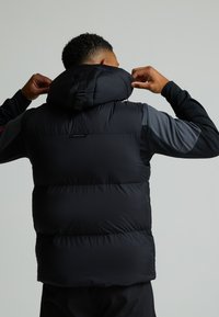 Μαύρο αμάνικο μπουφάν με ψηλό γιακά και ρυθμιζόμενη κουκούλα. Διαθέτει λεία υφή, οριζόντιο ραμμένο σχέδιο και ένα μικρό λογότυπο στην πλάτη.