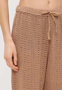 Κοντινή λήψη της μέσης ενός ατόμου και του χεριού που φορά μπεζ βελονάκι παντελόνι με κορδόνι που δένει στη μέση και ορατό αφαλό.