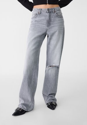 Jean gris clair à jambes larges avec un genou déchiré porté avec des chaussures pointues noires avec des boucles et œillets argentés.