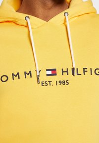 Sudadera amarilla con capucha de algodón, con un logo frontal "TOMMY HILFIGER EST. 1985" en azul marino, con cordones blancos y detalles metálicos.