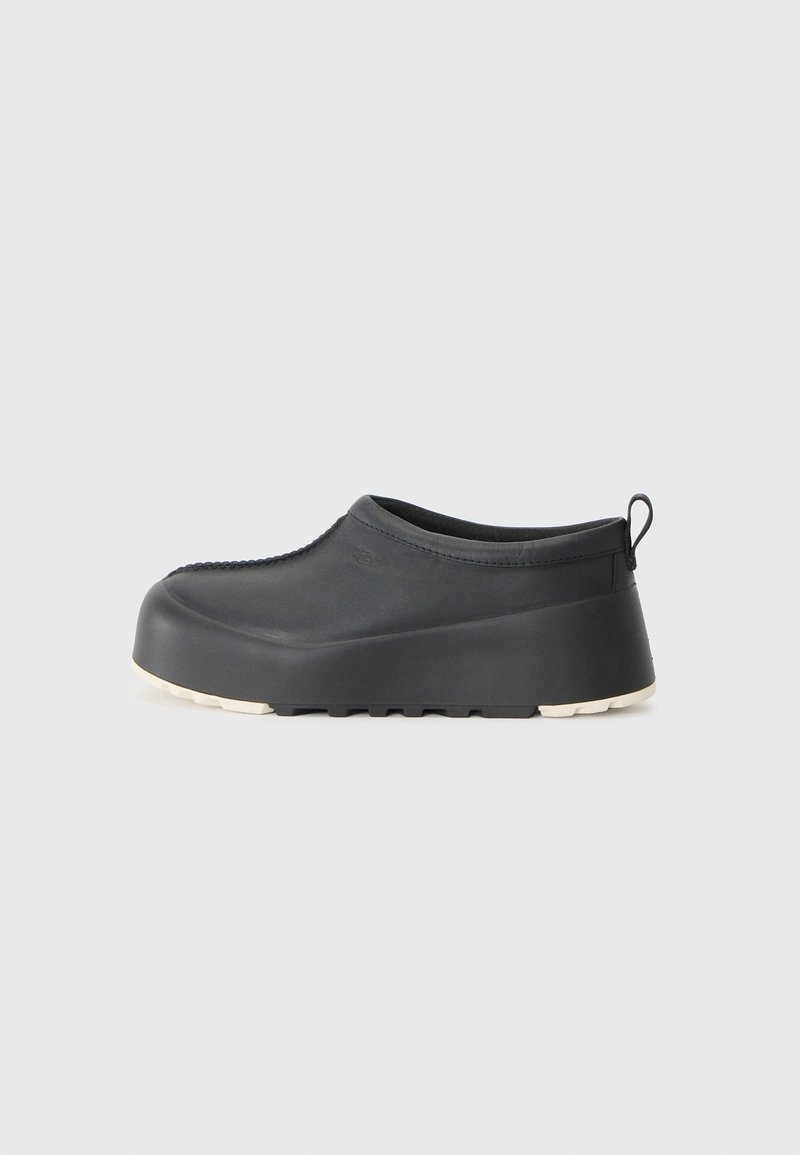 Fekete slip-on cipő sima gumiból, kerek orral, kiemelkedő sarkal és fehér texturált talppal. Hátul hurokkal a könnyű felhúzás érdekében.