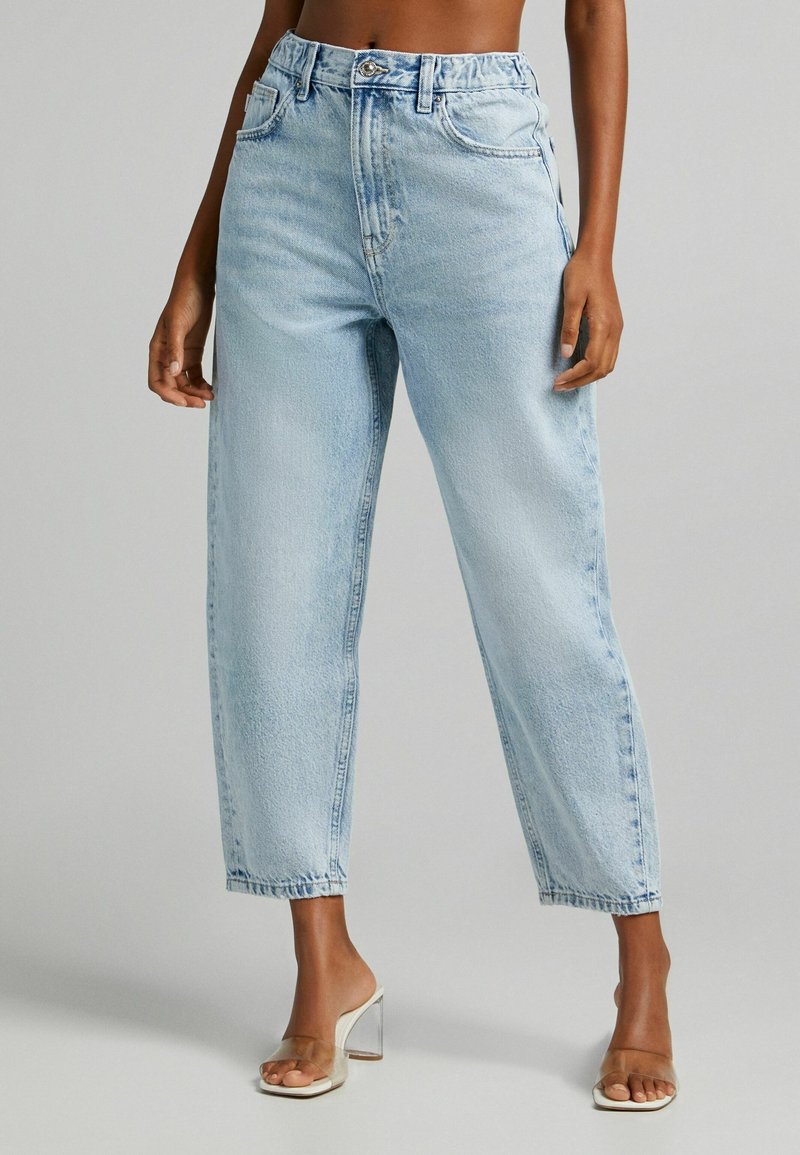 Light blue high-waisted jeans in a relaxed fit, featuring five pockets, subtle fading, and rolled hems paired with clear heeled sandals.