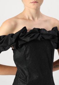 Μαύρο αμάνικο φόρεμα με γυαλιστερό ύφασμα, μεγάλη υφασμάτινη ρίγα κατά μήκος του άνω περιθωρίου και εφαρμοστή σιλουέτα με διακριτική λάμψη.