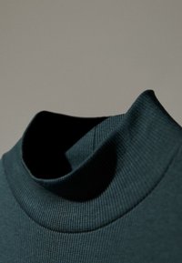 Mörk teal polotröja med ribbad struktur. Kragen är bred och strukturerad, vilket ger en tät passform runt halsen. Slät tyg.