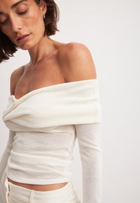 Off-shoulder, langærmet top i lys cremefarvet stof med samlet tekstur og ribbet detalje, tætsiddende design, blød og let gennemsigtig udseende.
