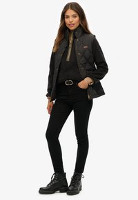 Zwarte gestikte vest over een donkere sweatshirt, zwarte jeans en zwarte veterschoenen. Heeft gouden accenten en een ritsdetail.