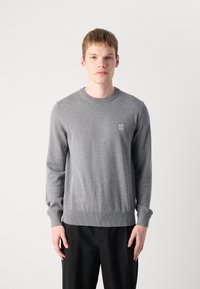 Junger Mann in einem schmal geschnittenen grauen Rundhalsausschnitt-Pullover mit einem kleinen quadratischen Logo auf der Brust, kombiniert mit schwarzen Hosen, stehend vor einem schlichten Hintergrund.