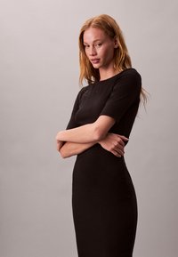 Tânără cu păr roșcat lung, purtând o rochie neagră mulată cu mâneci până la cot, stând cu brațele încrucișate pe un fundal simplu.