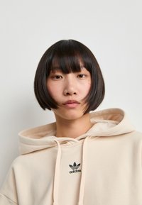 Sweatshirt bege com cordão ajustável e logotipo da Adidas em preto bordado. Textura suave, ajuste oversized, com um capuz grande e confortável.