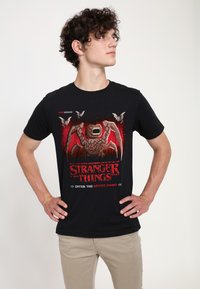 Camiseta negra de algodón con un diseño gráfico que muestra una criatura, murciélagos y el texto «Stranger Things» en rojo, sobre un fondo texturizado.