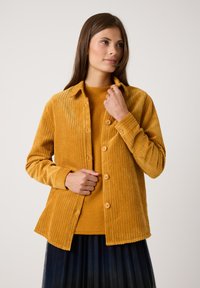 Senfgelber Cordjacke mit Knopfverschluss, taillierter Passform und sichtbarer Rippstruktur. Getragen mit einem passenden Strickoberteil.