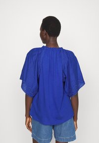 Blusa blu con maniche trasparenti ricamate e scollatura arricciata. Tessuto morbido, vestibilità rilassata, pieghe posteriori e bordi delle maniche a scallop.