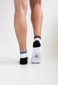 Schwarz-weiße Socken mit einem gestreiften Muster im oberen Bereich. Der untere Teil ist einfarbig weiß mit Logo-Detail auf der Rückseite. Aus Baumwollmaterial.