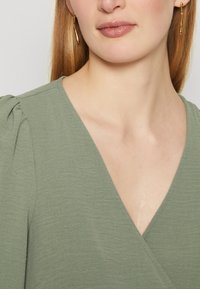 Olive green wrap top with a V-neckline, textured fabric, and soft pleats at the shoulders. Gold elongated hoop earrings.