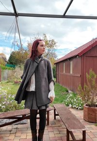 Capa cinzenta texturada com gola alta e fecho decorativo, combinada com uma blusa branca e uma saia aos quadrados cinzenta, ambientada num jardim.