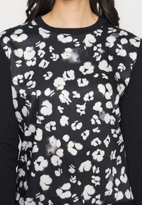 Žena v černo-bílém dlouhém rukávu s abstraktním bílým květinovým vzorem na přední straně a jednobarevnými černými rukávy.