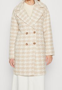 Beige och vit houndstooth-kappa med dubbelknäppt design, stora slag, två bruna knappar och en texturerad tyg.