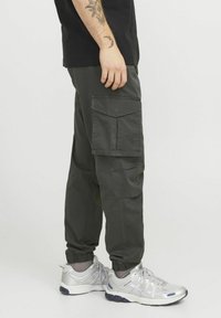 Mørkegrønne cargo bukser med flere lommer, elastiske manchetter og en løs pasform, parret med sølvfarvede sneakers og en sort t-shirt.
