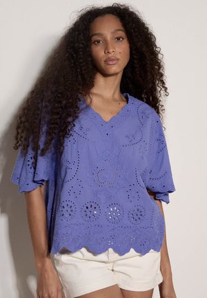 Kobieta z długimi kręconymi włosami, ubrana w luźną fioletową bluzkę z haftem ażurowym oraz białe szorty, stojąca na jasnym tle.