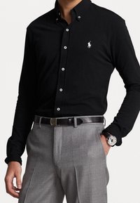 Hombre vestido con camisa de botones negra de manga larga con un logo blanco, pantalones grises, cinturón de cuero negro, reloj de pulsera, posando con una mano en el bolsillo.