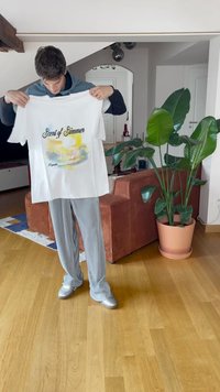 Fehér pamut póló, amelyen a "Nyár Illata" felirat és egy akvarell tervezés található. A modell a pólót tartja, világosszürke csíkos nadrággal és ezüst színű cipővel párosítva.