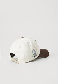 Hvid baseballkappe med en brun skygge, prydet med et broderet mærke på bagsiden, der fremhæver 1983 og 40 år. Justerbar rem bagpå.