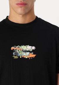 Tricou negru din bumbac, având un grafic colorat al unui monstru stilizat cu detalii verzi, portocalii și albe. Guler rotund, maneci scurte.