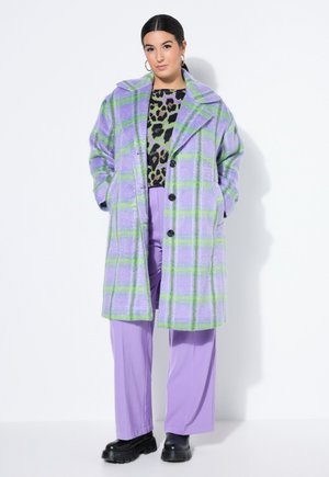 Kvinde iført en lilla og grøn ternet frakke over en leopardmønstret top, lilla bukser og sorte sko med chunky sål, stående op ad en hvid væg.