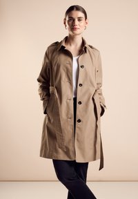 Trench beige con cierre de botones, hombreras, y bolsillos laterales, que presenta un cinturón atado y un corte relajado.