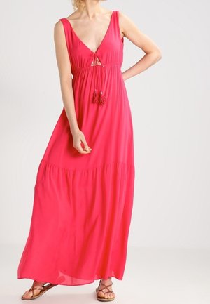 Kobieta ubrana w jasnoróżową, bezrękawową suknię maxi z głębokim dekoltem w kształcie litery V i zawiązywaną frędzlami, w połączeniu z brązowymi płaskimi sandałami.