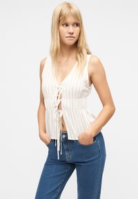 Νεαρή γυναίκα που φοράει λευκή ριγέ αμάνικη μπλούζα με μπροστινούς δεσμούς και μπλε τζιν, στέκεται με τα χέρια στις τσέπες, κοιτώντας στο πλάι.