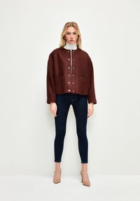 Braune, strukturierte, kurz geschnittene Jacke mit vorderer Knopfleiste und zwei Taschen, kombiniert mit dunklen, engen Jeans und beigen Absatzschuhen. Darunter ein weißer Rollkragenpullover.