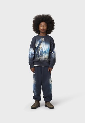 Kind mit großem Afro, trägt dunkelblauen Dinosaurier-Sweatshirt und passende Hose, steht mit neutralem Gesichtsausdruck und braunen Stiefeln auf weißem Hintergrund.